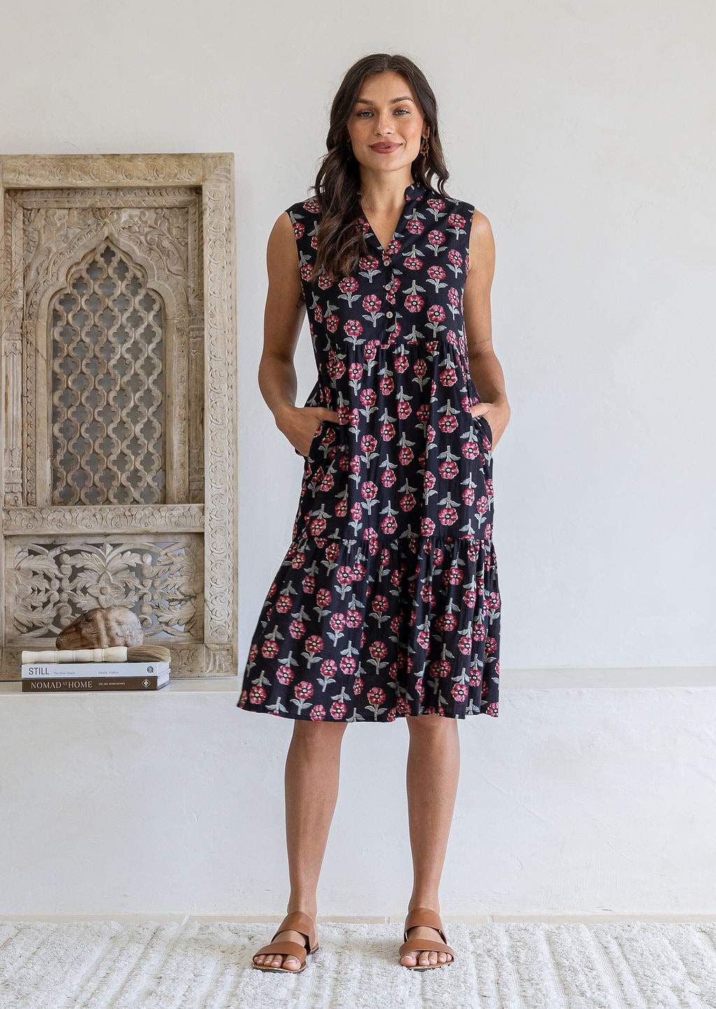 Woman wearing a knee length cotton sleeveless dress with buttoned bodice and  two tiered skirt inn earthy black based floral print.