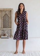 Woman wearing a knee length cotton sleeveless dress with buttoned bodice and  two tiered skirt inn earthy black based floral print.
