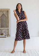 A woman modelling a sleeveless black based floral print cotton midi dress with a one hand in pockets
