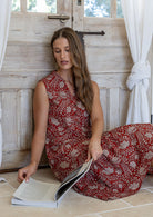 Woman in a red floral dress reading a book in a sunlit room with wooden walls.