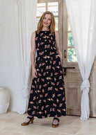 Woman wearing a cotton black based floral sleeveless maxi dress standing in a room with white curtains and wooden door.