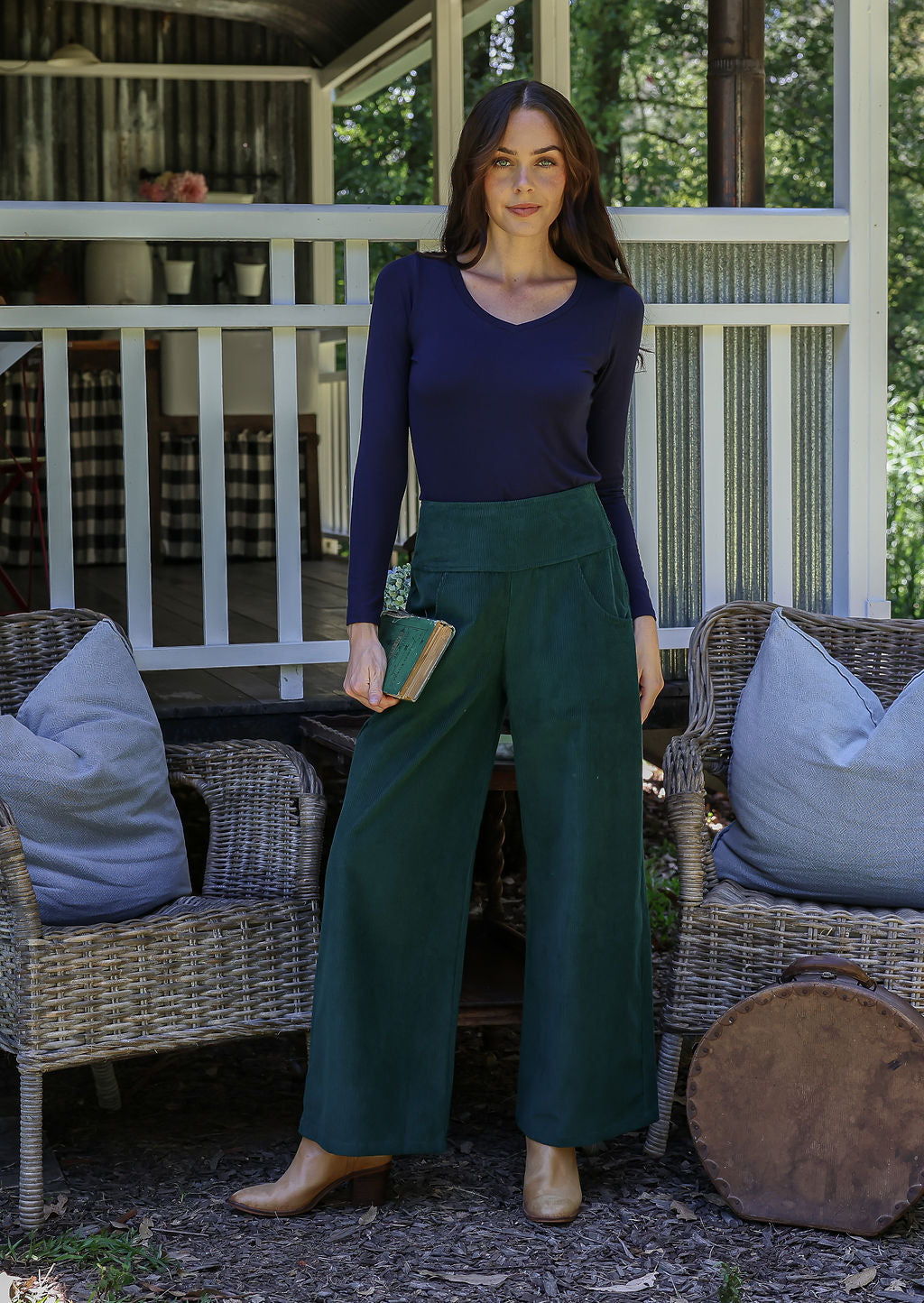 Woman wearing a navy blue top and green corduroy pants standing on a porch with wicker furniture and cushions.