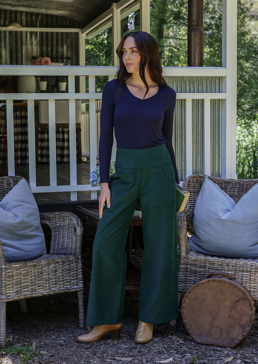 Woman standing on a porch wearing a dark blue top and green pants.