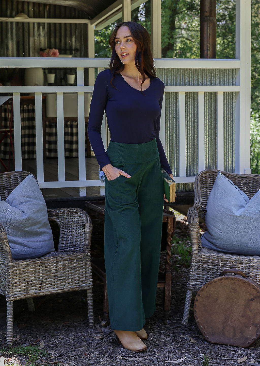 Woman in a navy top and green pants standing on a porch with one hand in pocket