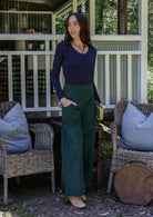Woman in a navy top and green pants standing on a porch with one hand in pocket