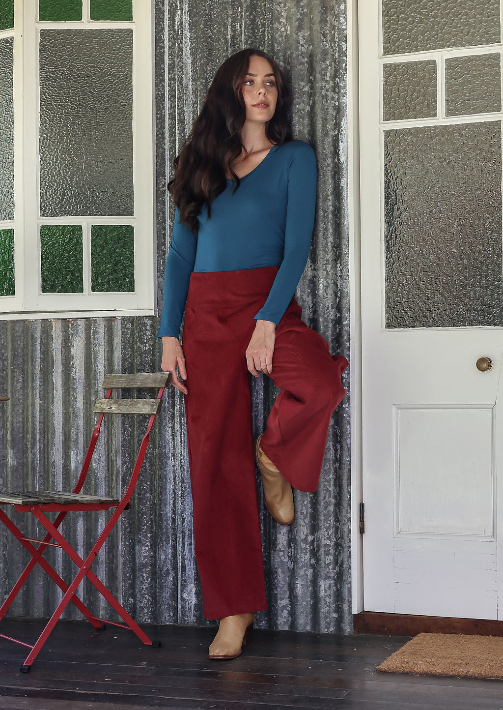 Woman wearing a cotton corduroy pants in cabernet standing in a room with a red chair and patterned wallpaper.