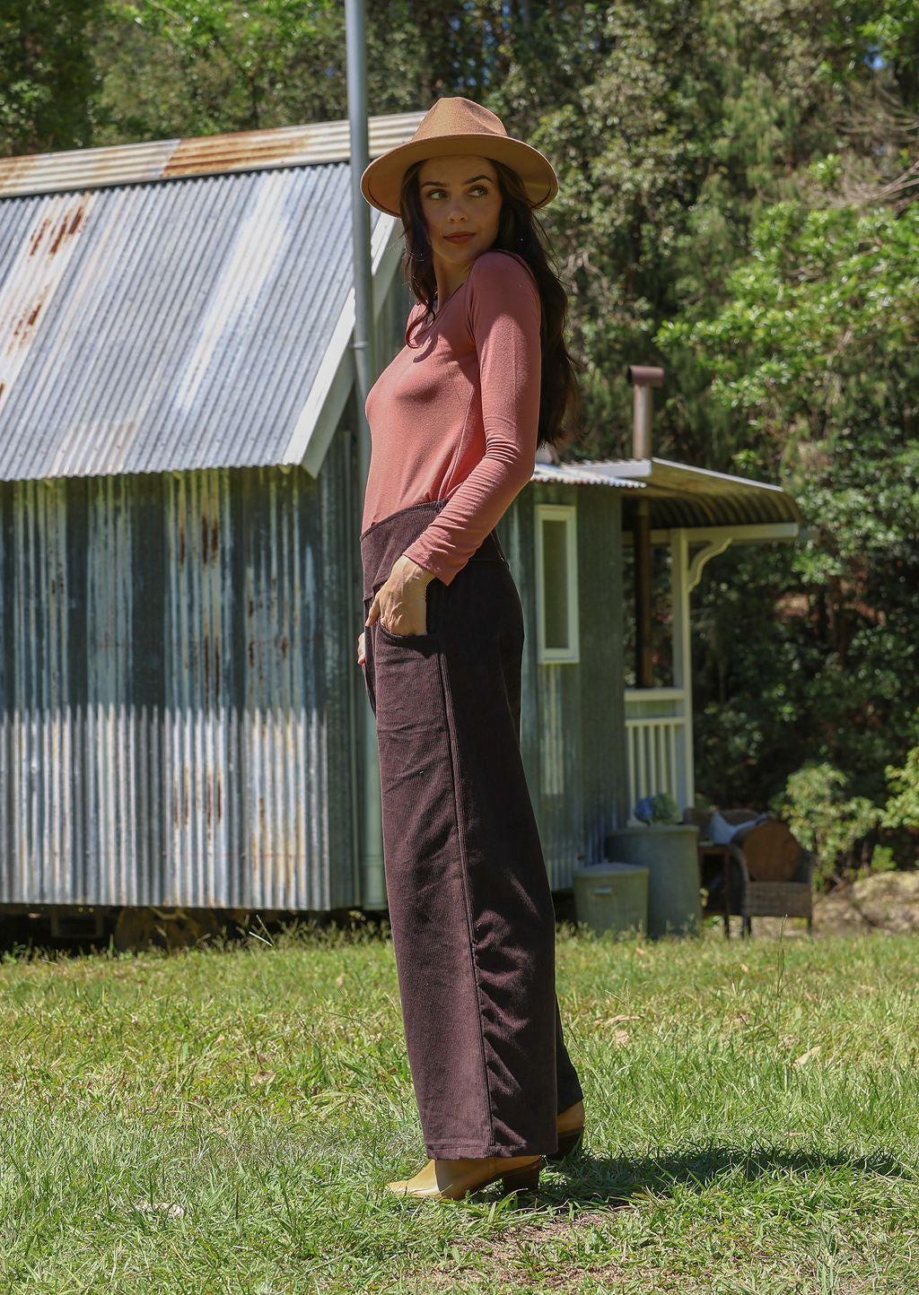 Woman in a pink top and brown corduroy pants standing in front of a rustic cabin.