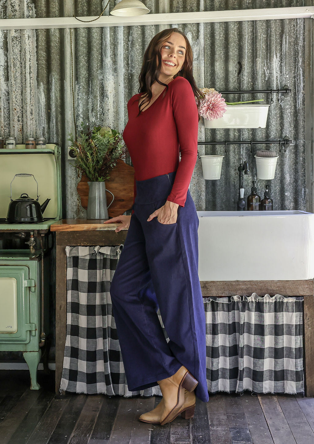 Woman in a red top and blue cotton corduroy pants standing in a rustic kitchen with hand in pocket