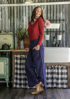Woman in a red top and blue cotton corduroy pants standing in a rustic kitchen with hand in pocket