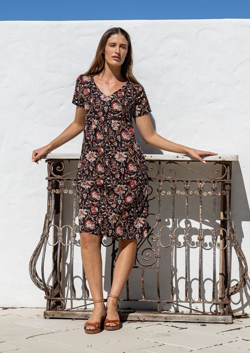 Woman in a floral dress standing against a white wall with a decorative metal railing.