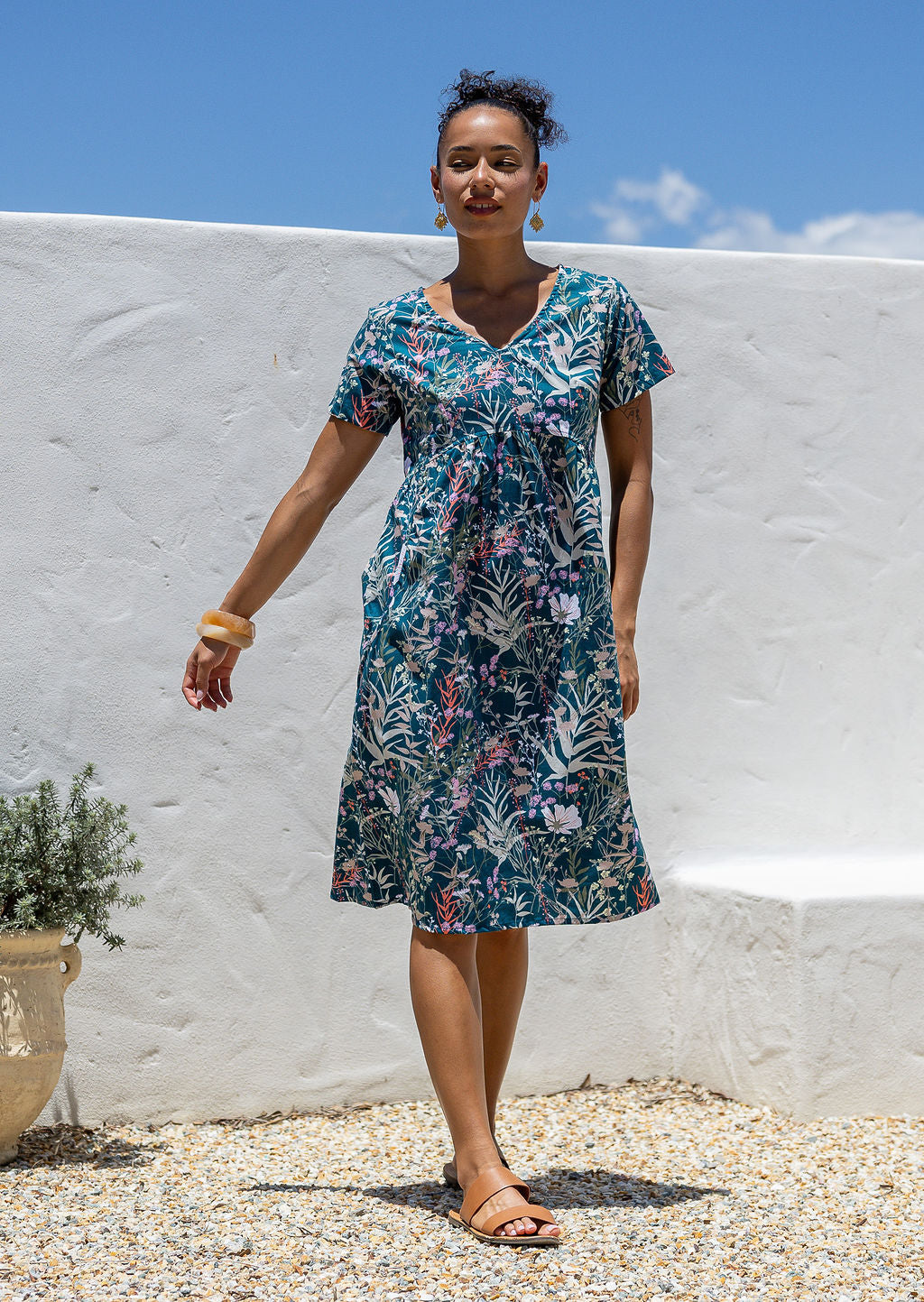 Woman wearing a teal cotton floral dress with v-neck standing against a white wall with blue sky.