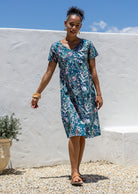 Woman wearing a teal knee length cotton floral dress with v-neck standing against a white wall with blue sky.