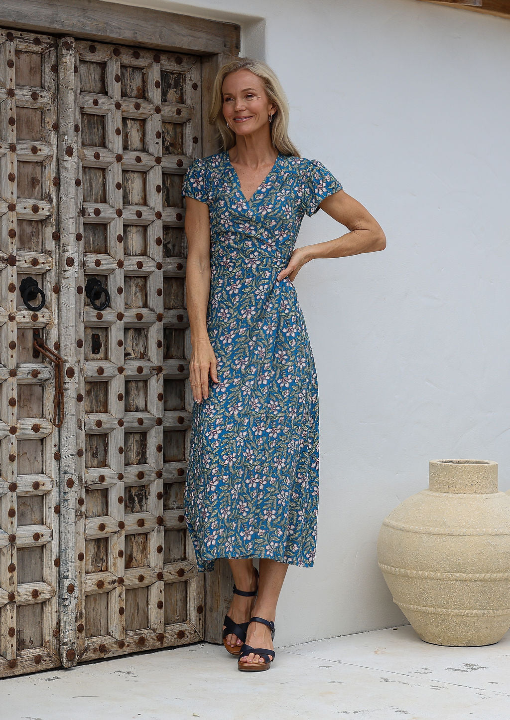 Woman in a blue floral rayon wrap dress with butterfly sleeves and small gathers at the shoulder, standing next to a wooden door.
