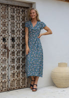 Woman in a blue floral rayon wrap dress with butterfly sleeves and small gathers at the shoulder, standing next to a wooden door.