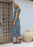 Woman in a blue floral wrap around maxi dress standing next to a textured wooden door and large beige vase.