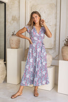 Woman wearing blue based floral print wrap around maxi dress standing against a white background