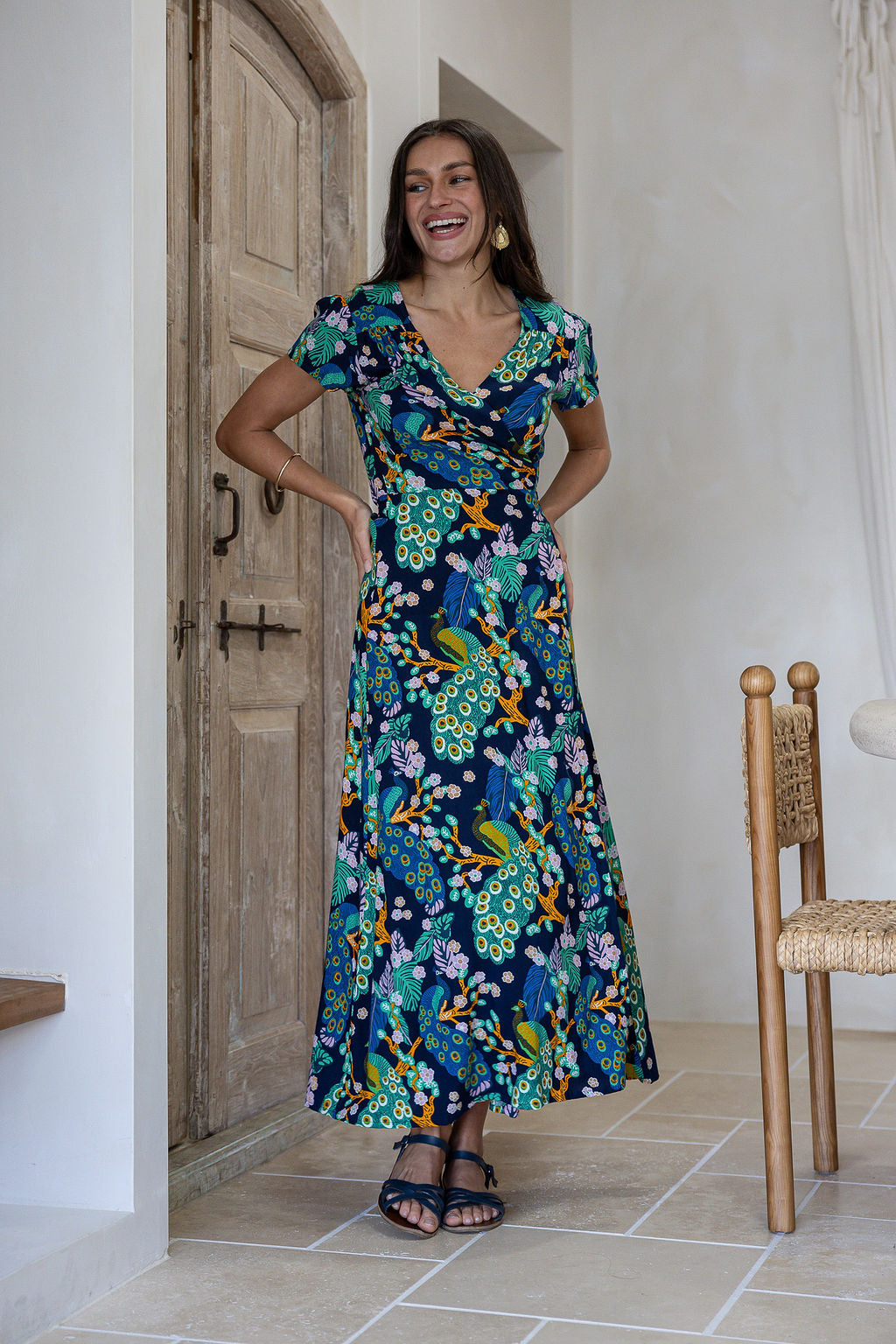Woman wearing a navy blue based floral print wrap around dress with butterfly sleeves, standing in front of a wooden door