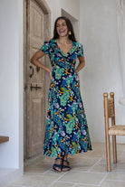 Woman wearing a navy blue based floral print wrap around dress with butterfly sleeves, standing in front of a wooden door