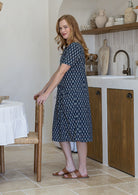Woman stands sideways wearing a 100% cotton dress in navy blue based floral print with short sleeves