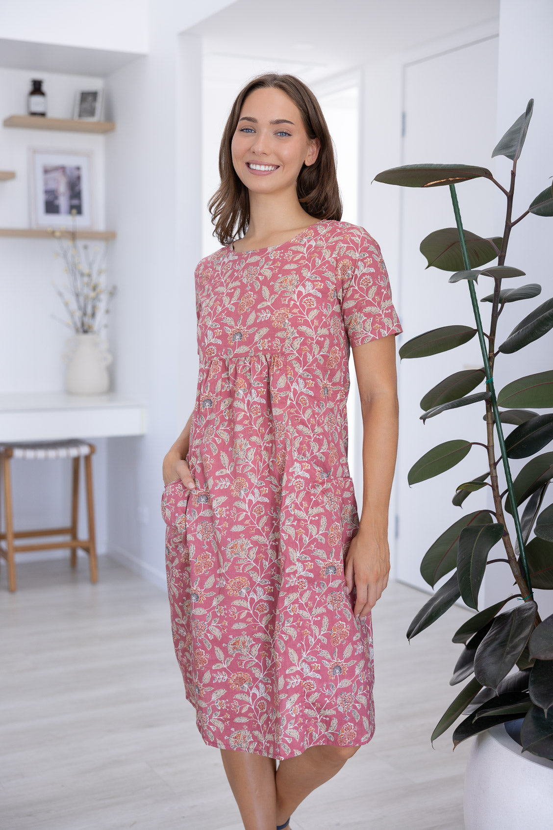 Woman wearing a 100^% cotton floral dress standing in a room with a plant and shelves.