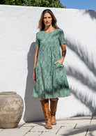 Woman wearing a light sage green cotton dress with a white wall and pot in the background