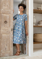 Woman in a blue floral dress standing in a doorway with shelves in the background