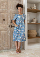 Woman wearing a blue floral cotton dress standing in a room with wooden doors and shelves.