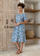 Woman wearing a blue floral dress standing in a room with wooden shelves and a wooden door.