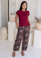 Woman wearing a red top and black based floral cotton pants standing in a decorative indoor setting with vases and plants.