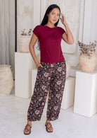 Woman standing in front of white wall wears 100% cotton wide leg Indian traditional print pants in black with elastic waist, pockets and pleated ankles