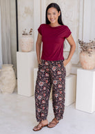 Woman wearing a maroon sleeveless top and floral wide leg cotton pants with hands in pockets, standing in front of a white wall