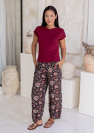 Woman wearing a maroon sleeveless top and black based floral print wide leg cotton pants with pleated ankles, elastic waist and pockets