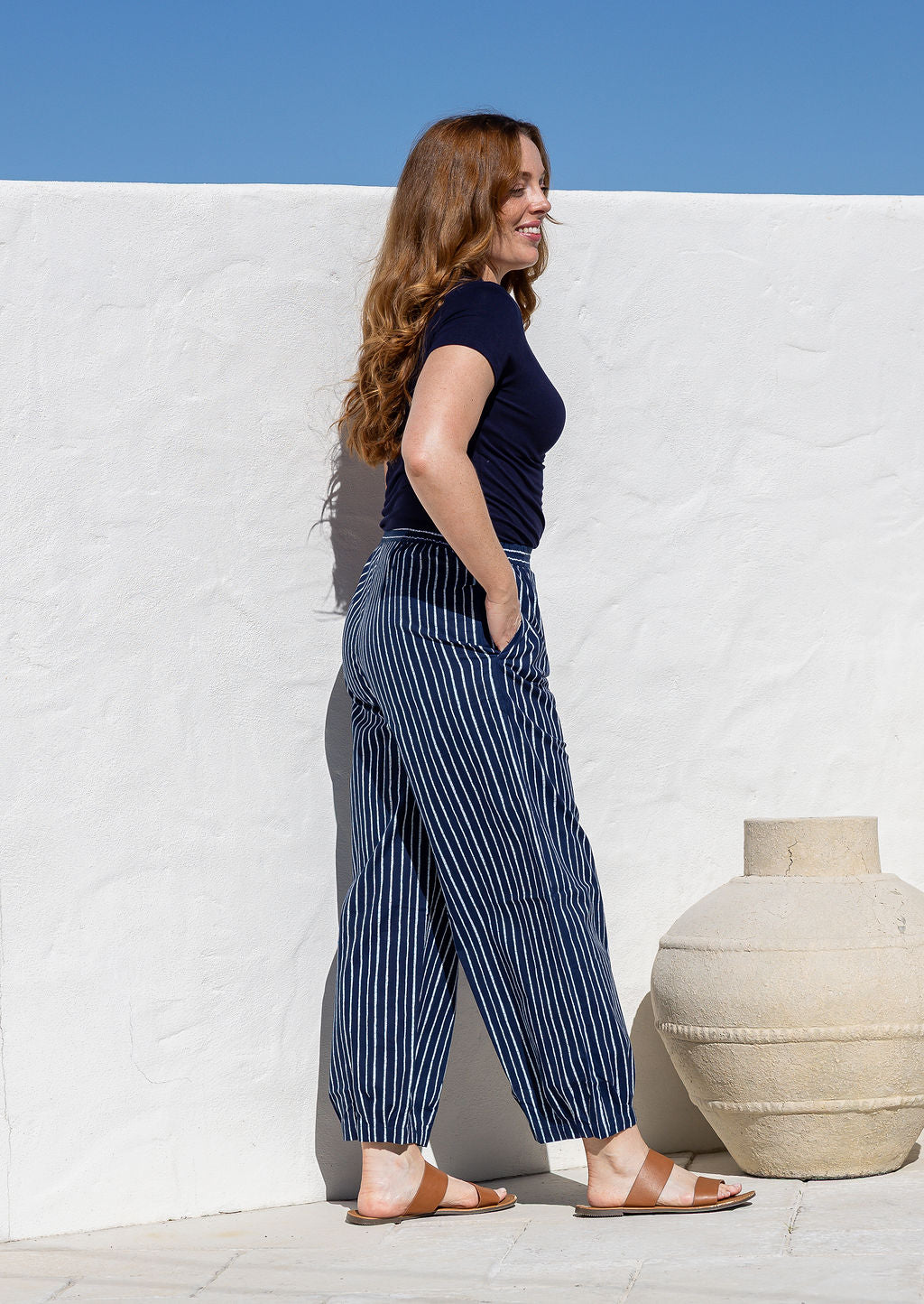 Woman stands sideways wearing a 100% cotton wide leg pants in navy based striped print with one hand in pocket 