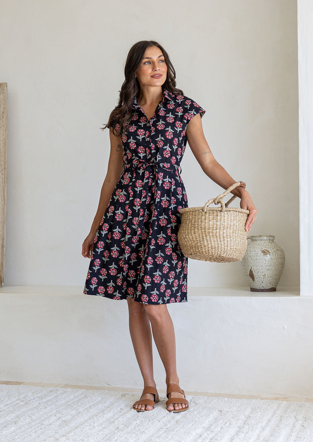 Woman wearing a 100% cotton black floral print shirt dress with tie waist with woven basket and sandals