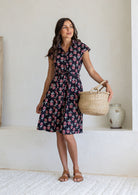 Woman wearing a 100% cotton black floral print shirt dress with tie waist with woven basket and sandals