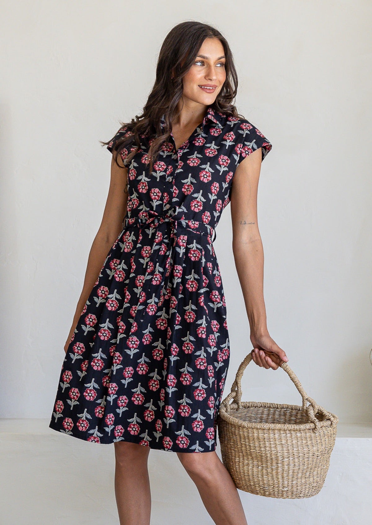 Woman wearing a 100% cotton black floral print Ursula Dress with cap sleeves and collar, holding woven basket.