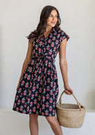 Woman wearing a 100% cotton black floral print Ursula Dress with cap sleeves and collar, holding woven basket.