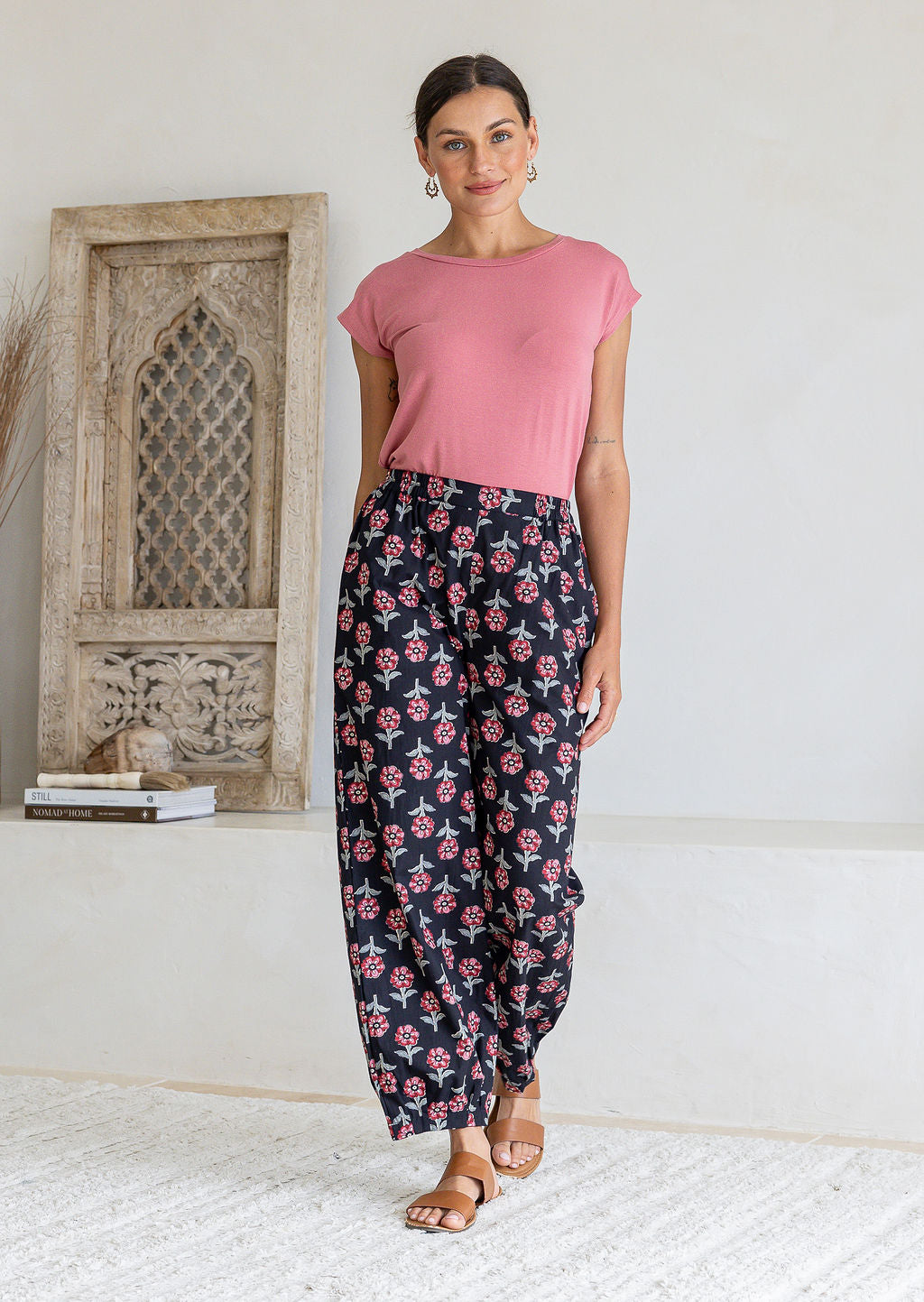 Model wearing black floral print wide-leg Greta Pants with a pink t-shirt and tan sandals. 
