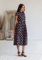 Woman stands sideways wearing a 100% cotton Black floral midi shirtdress with cap sleeves and tie waist. 