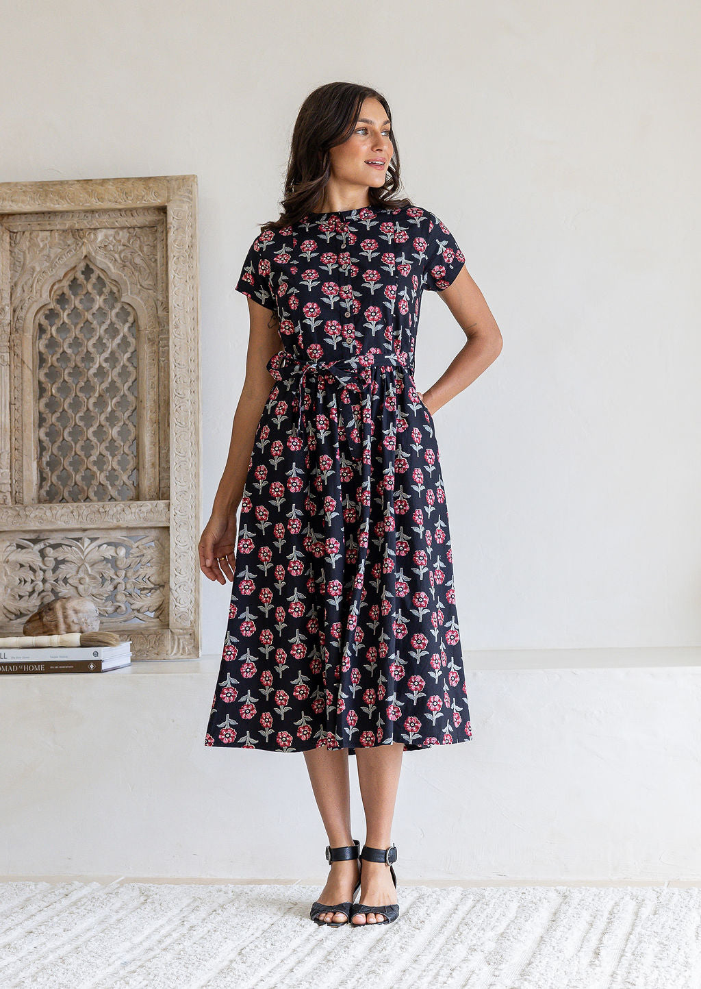 Woman with one hand in pocket of a black floral print midi dress with short sleeves and tie waist, 100% cotton. 