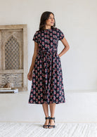 Woman with one hand in pocket of a black floral print midi dress with short sleeves and tie waist, 100% cotton. 
