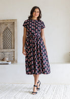 Woman poses in a pure cotton black floral print midi dress with short sleeves and gathered waist. 