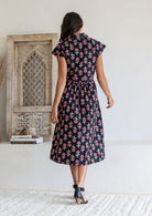 Woman stands backwards wearing a 100% cotton Black floral midi shirtdress with cap sleeves and tie waist. 