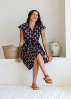 Woman wearing a black floral print wrap-style midi dress with cap sleeves, worn by model seated on ledge, tan sandals.