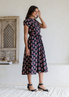 Model standing sideways wearing a black midi dress with floral print, short sleeves and tie waist. 