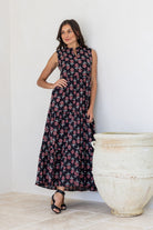 Black floral tiered maxi dress with sleeveless mandarin collar, worn by model standing against wall.
