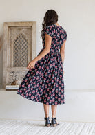Woman stands backwards wearing a 100% cotton black based floral print midi dress with cap sleeves paired with black heels.
