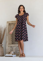 Woman poses in a 100% cotton black blue based floral dress with round neckline and cap sleeves
