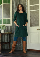 Woman wearing green corduroy A-line dress with 3/4 sleeves with hands in pockets over leggings and boots