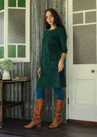 Woman in a green corduroy dress and brown knee-high boots standing in a room with patterned walls.